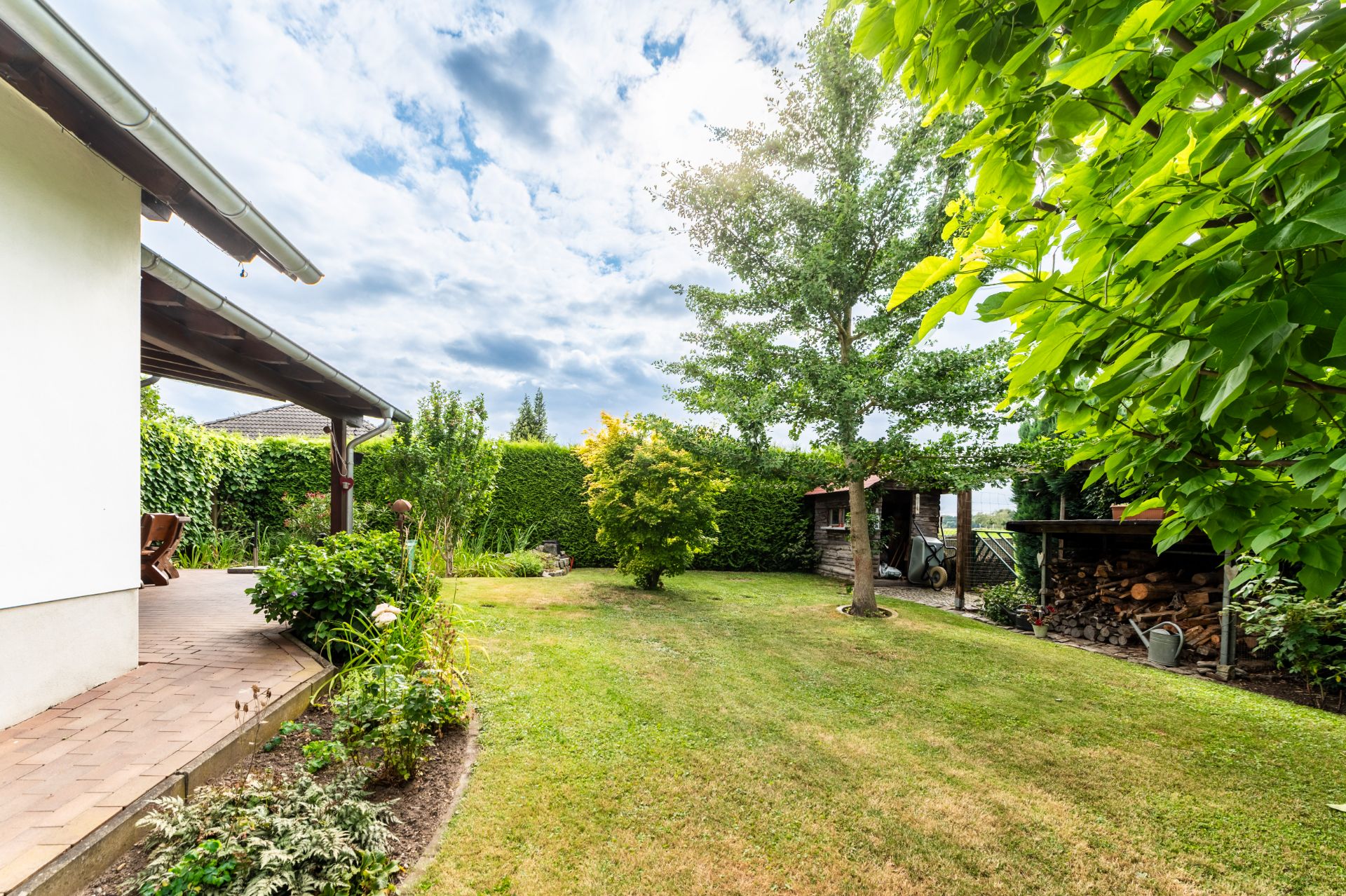 Garten mit Rasen, Bäumen, Terrasse und Holzlager an einem Wohnhaus bei Tageslicht.