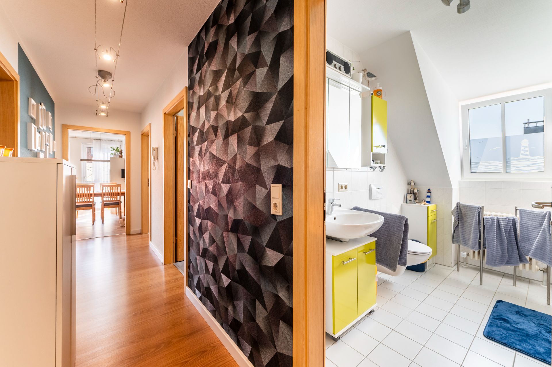 Flur mit Holzboden, gemusterter Wand und Blick ins helle Badezimmer sowie Esszimmer.