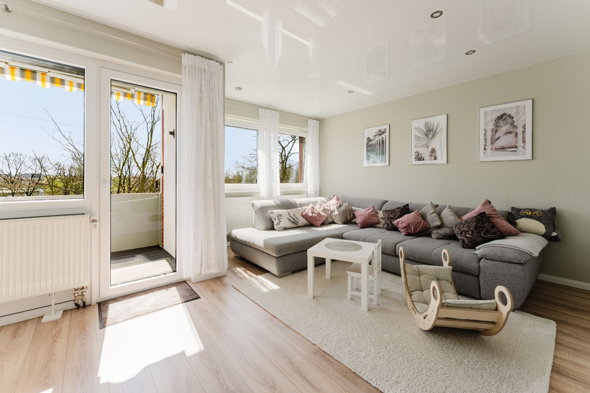 Helles Wohnzimmer mit Ecksofa, Couchtisch, Kinderstuhl und Zugang zum Balkon.