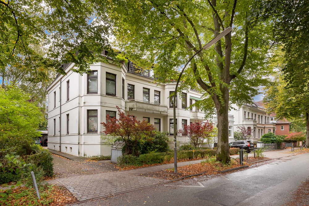Weiße Wohnhäuser an einer von Bäumen gesäumten Straße mit Gehweg und parkenden Autos.