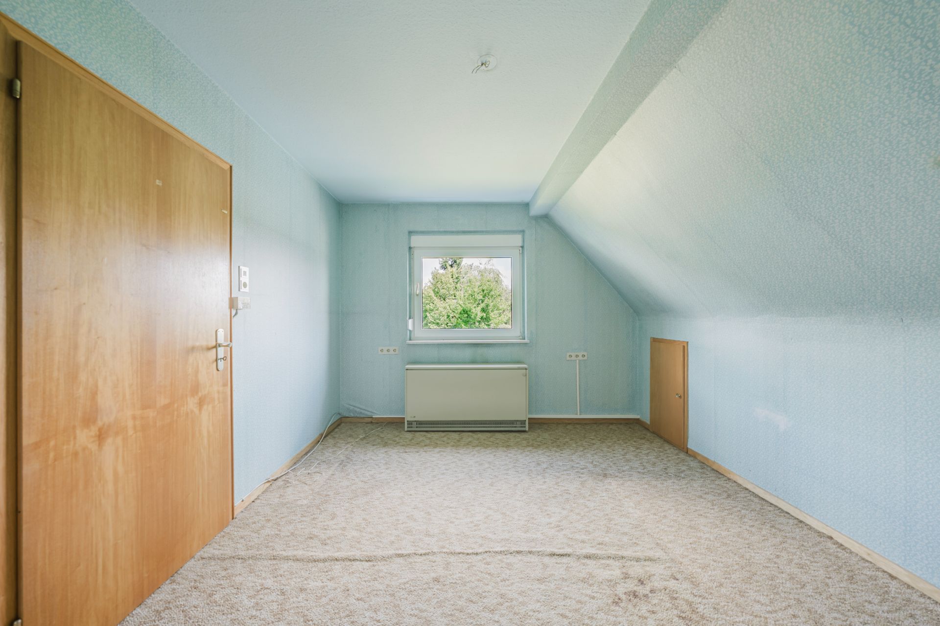 Leeres Zimmer mit blau gestrichenen Wänden, Teppichboden, Holztür und Fenster zur Außenseite.