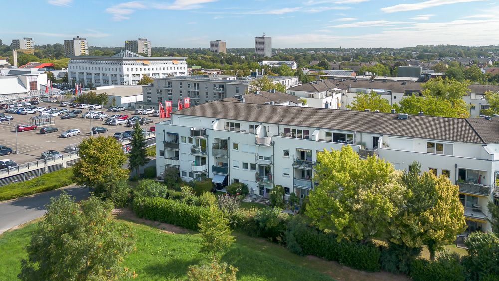 Mehrfamilienhaus mit Balkonen und umliegenden Bäumen, im Hintergrund Parkplätze und weitere Gebäude.
