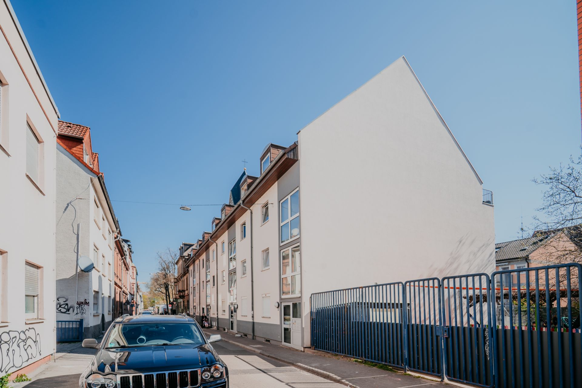 Wohnstraße mit mehrstöckigen Häusern, schwarzem Auto und blauem Zaun bei Tageslicht.