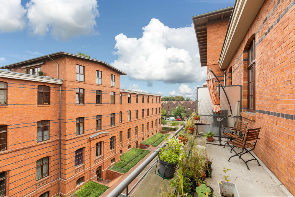 Balkon mit Pflanzen und Sitzgelegenheit, Blick auf roten Backsteinbau und Himmel mit Wolken.