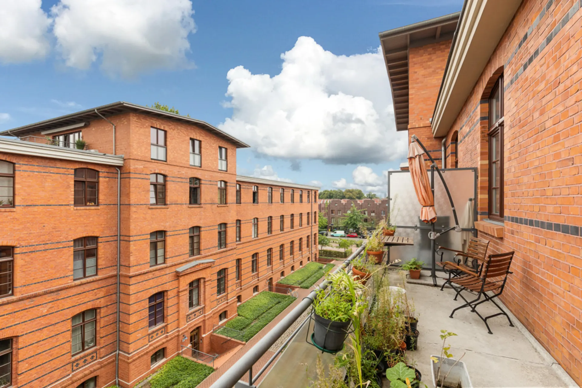 Balkon mit Pflanzen und Sitzgelegenheit, Blick auf roten Backsteinbau und Himmel mit Wolken.