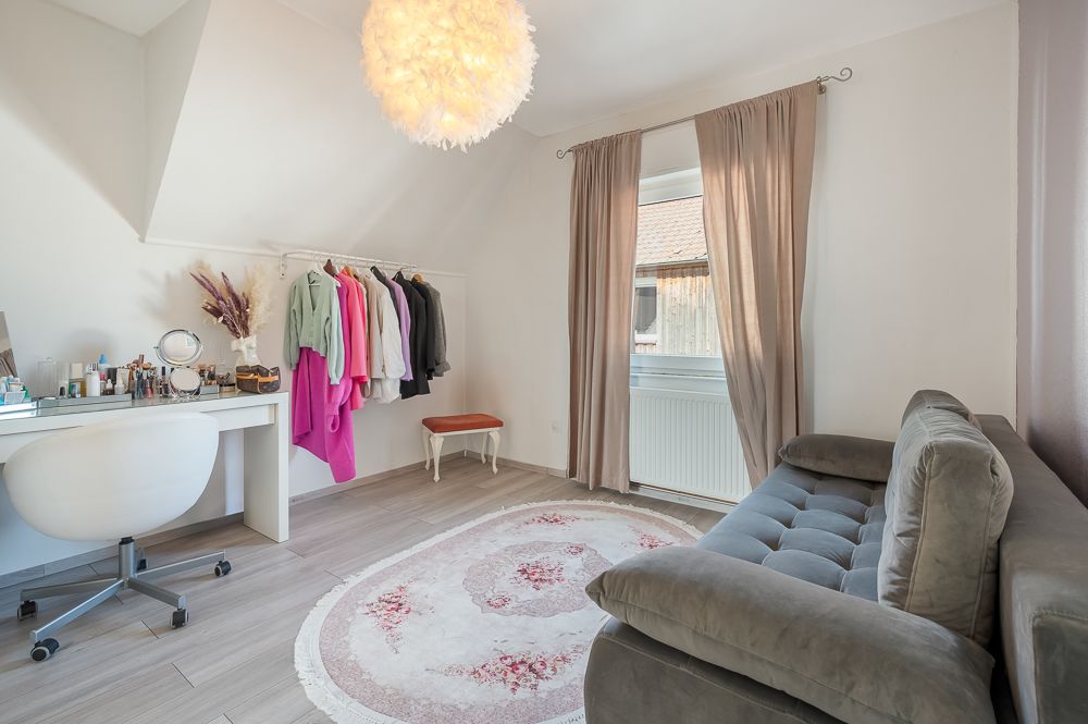 Helles Zimmer mit grauem Sofa, Schminktisch, Kleiderständer und rundem Teppich.
