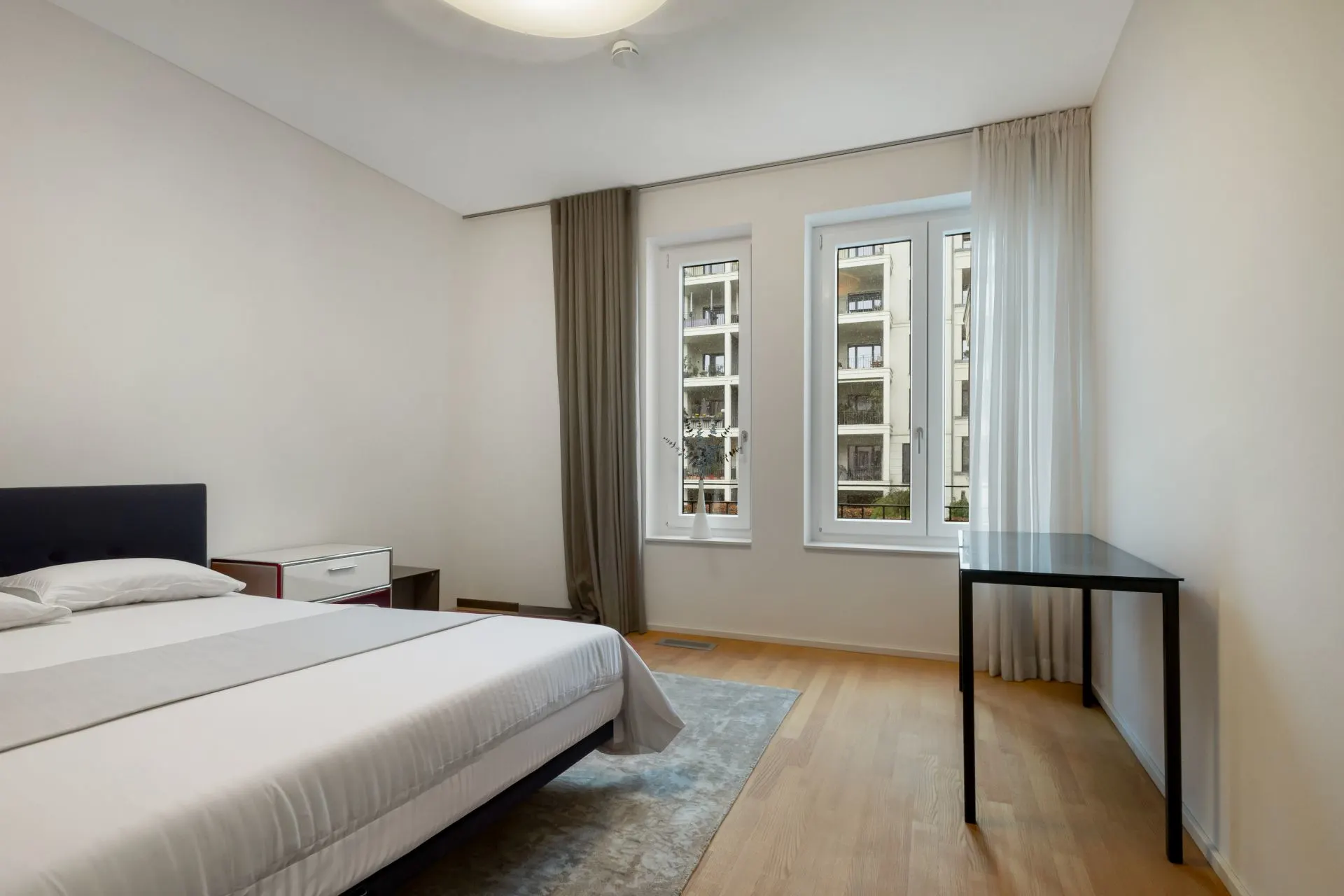 Helles Schlafzimmer mit Bett, Nachttisch, Schreibtisch und zwei großen Fenstern.