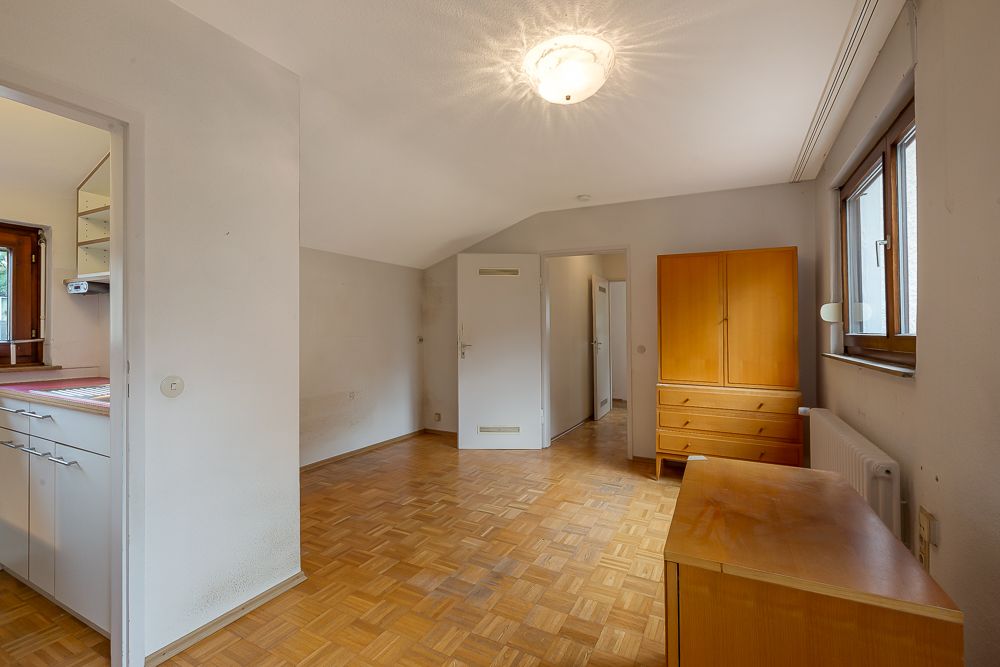 Helles Zimmer mit Parkettboden, Holzschrank, Kommode und Fenster, Küche und Flur sichtbar.