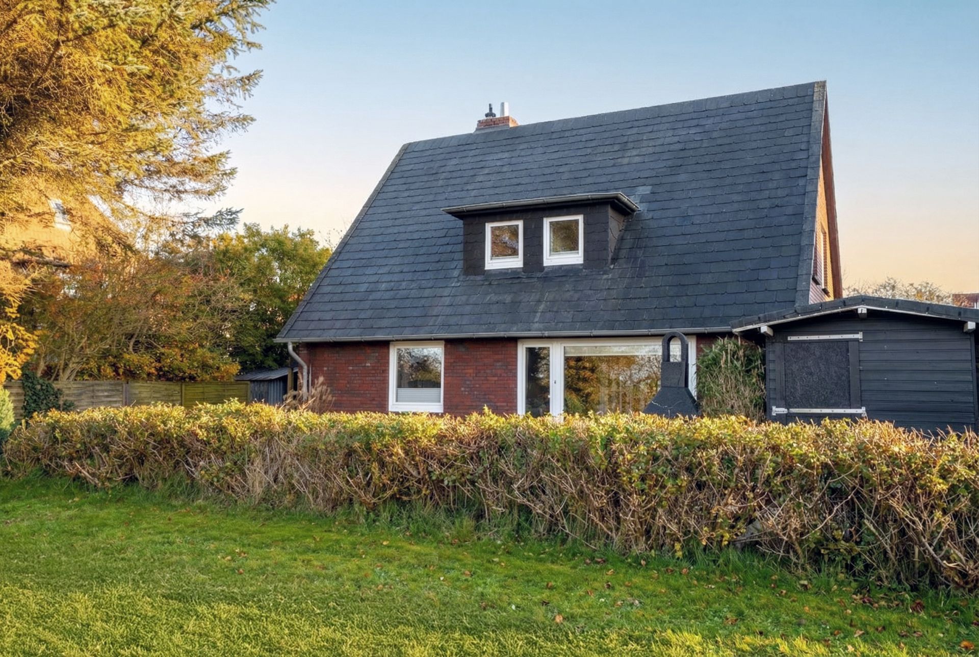 Haus mit dunklem Satteldach, roter Fassade und großem Fenster, umgeben von Garten und Hecke.