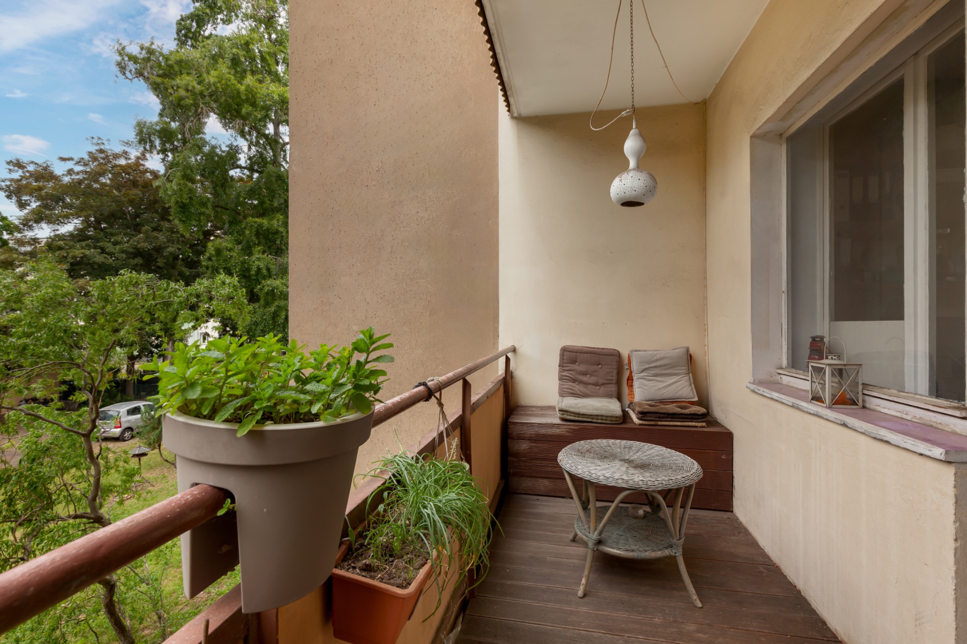Balkon mit Sitzbank, kleinem Tisch, Pflanzen und Blick auf grüne Bäume.