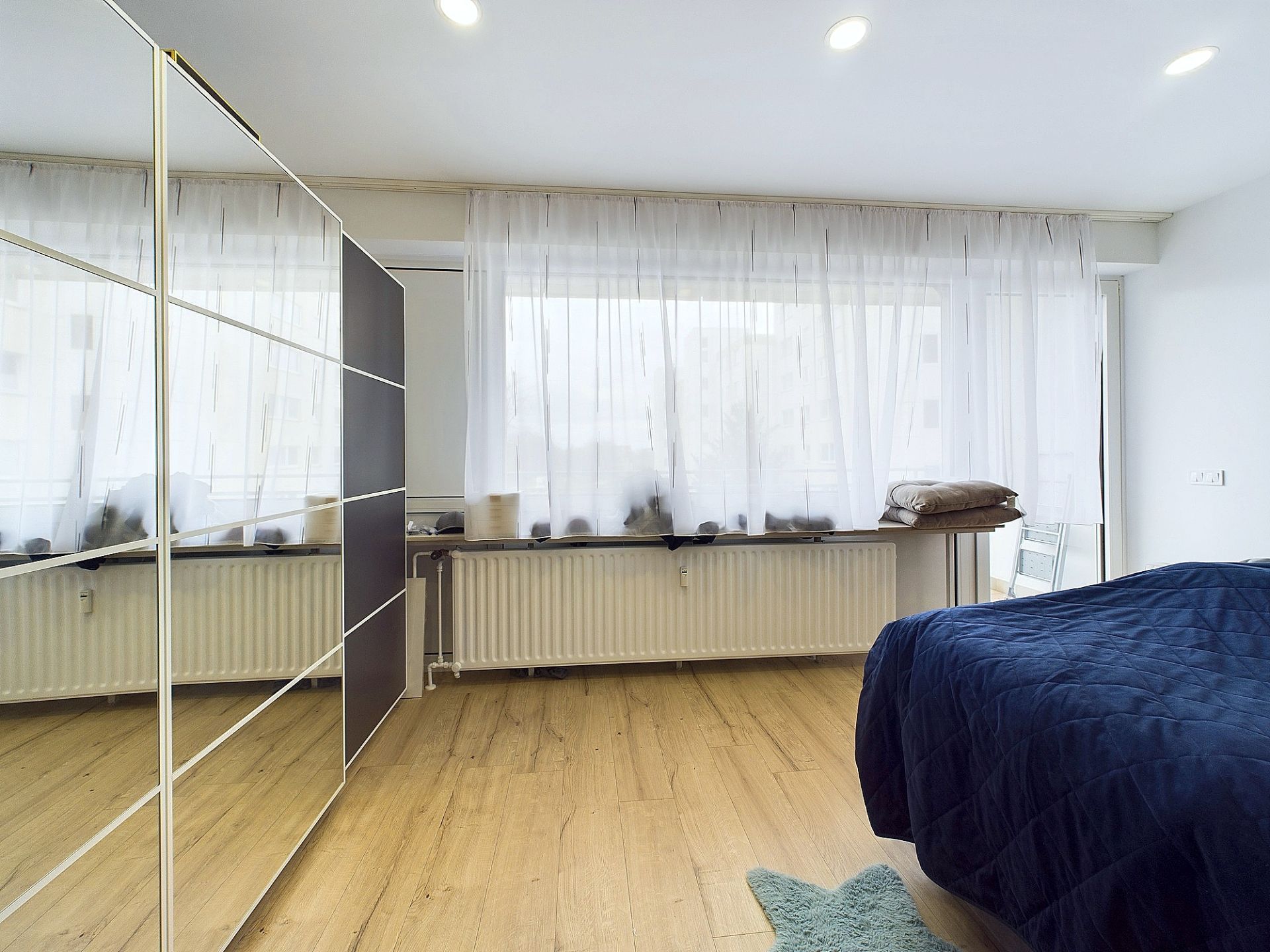Helles Schlafzimmer mit großem Fenster, Holzboden, Spiegelkleiderschrank und blauem Bett.