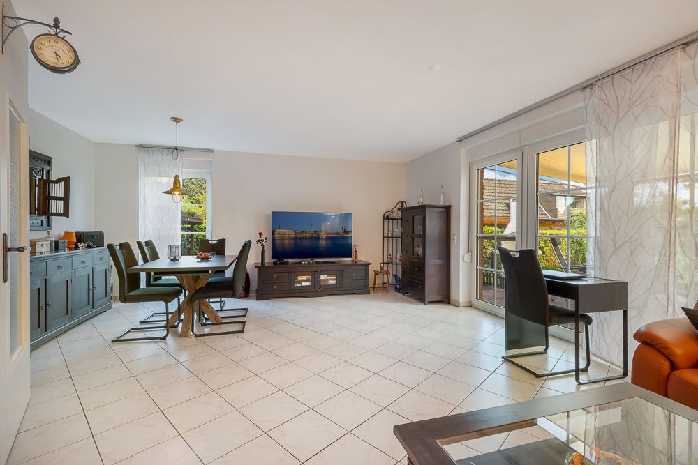 Helles Wohnzimmer mit Esstisch, Fernseher, Schreibtisch und großen Fenstern zum Balkon.