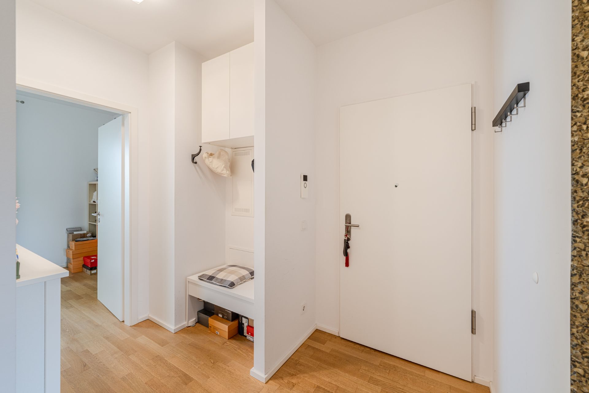 Heller Flur mit Holzfußboden, weißer Tür, Garderobe und Sitzbank mit Kissen.