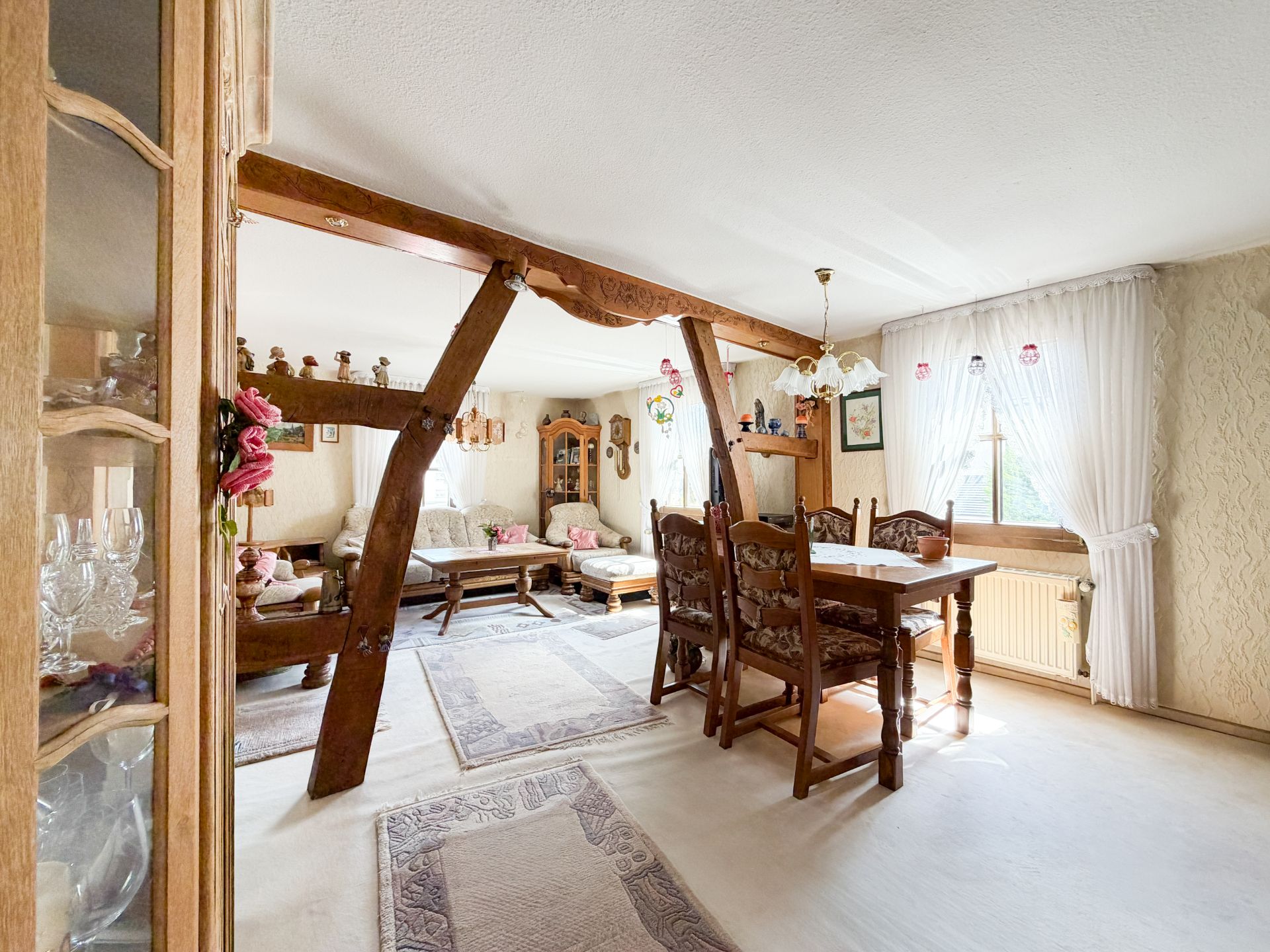 Wohnzimmer mit Holzbalken, Esstisch, Stühlen, Sofa und großen Fenstern mit Gardinen.