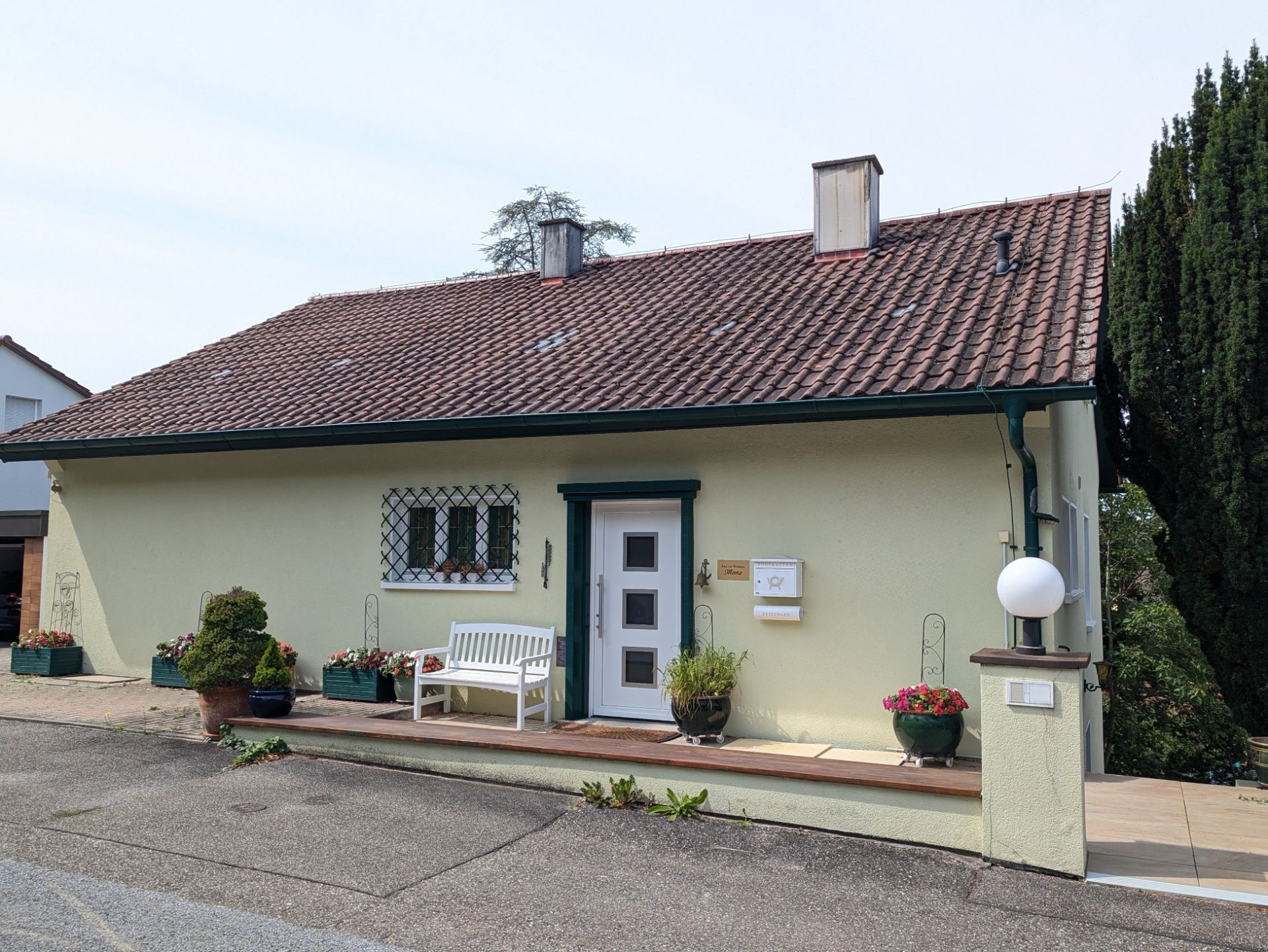 Gelbes Einfamilienhaus mit rotem Dach, weißer Bank und Blumenkübeln vor dem Eingang.