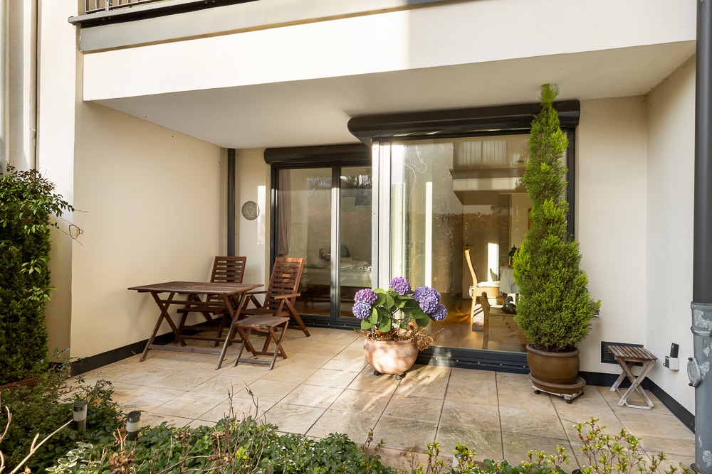Überdachte Terrasse mit Holztisch, Stühlen, Blumenkübeln und Glasschiebetür zum Wohnbereich.