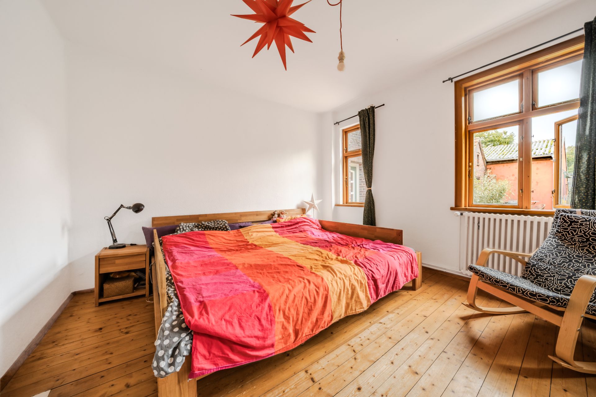 Schlafzimmer mit Holzboden, Doppelbett, Nachttisch, Sessel und zwei Fenstern mit dunklen Vorhängen.
