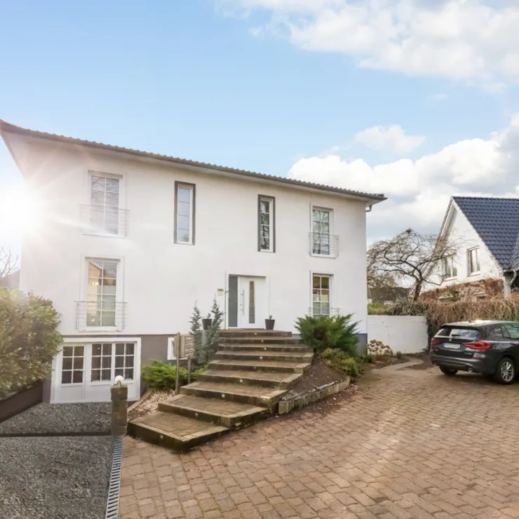Weißes zweistöckiges Wohnhaus mit Treppe zum Eingang und geparkten Autos auf dem Hof.