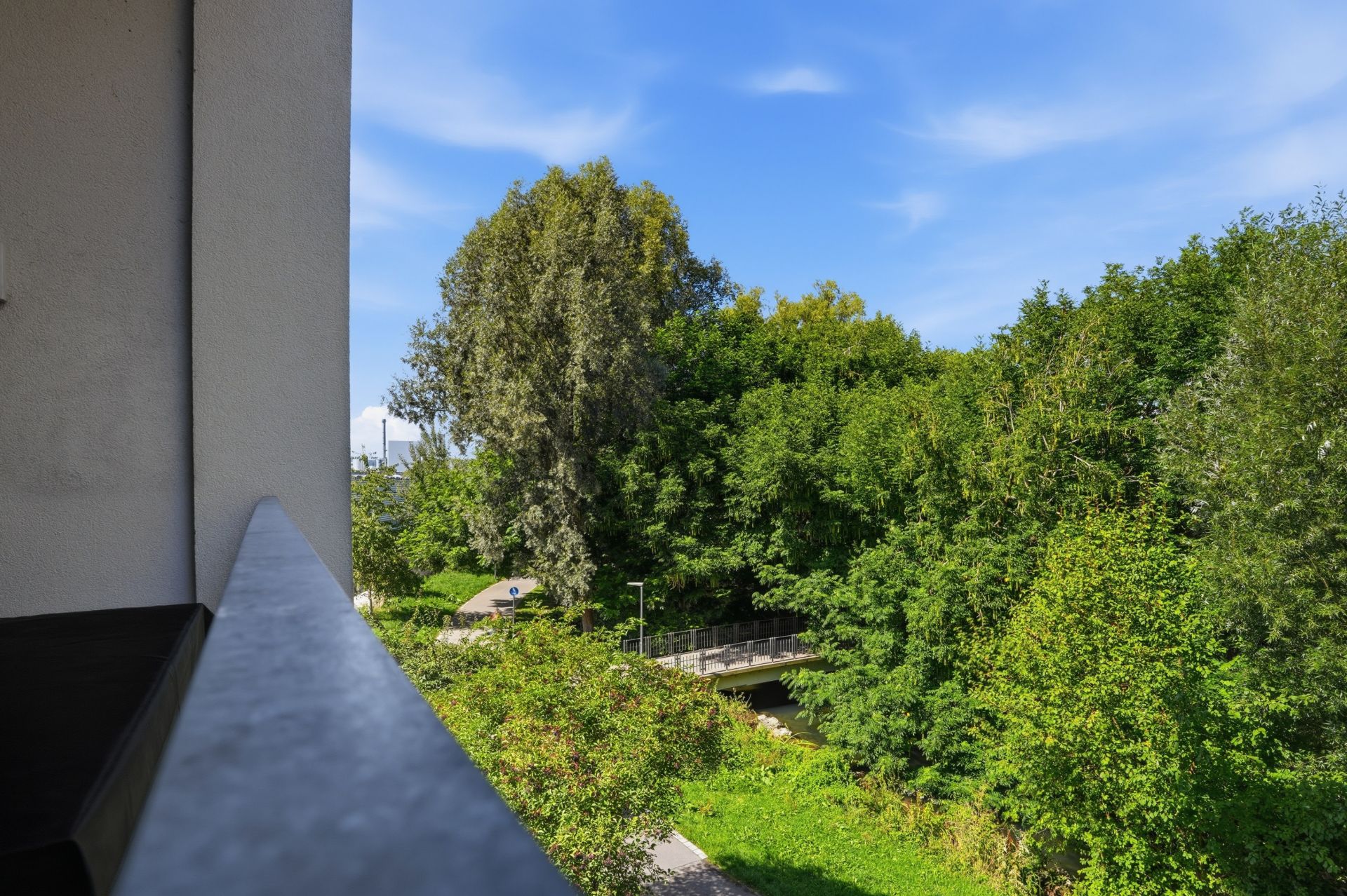 Blick von einem Balkon auf eine grüne Baumlandschaft und einen Weg unter blauem Himmel.