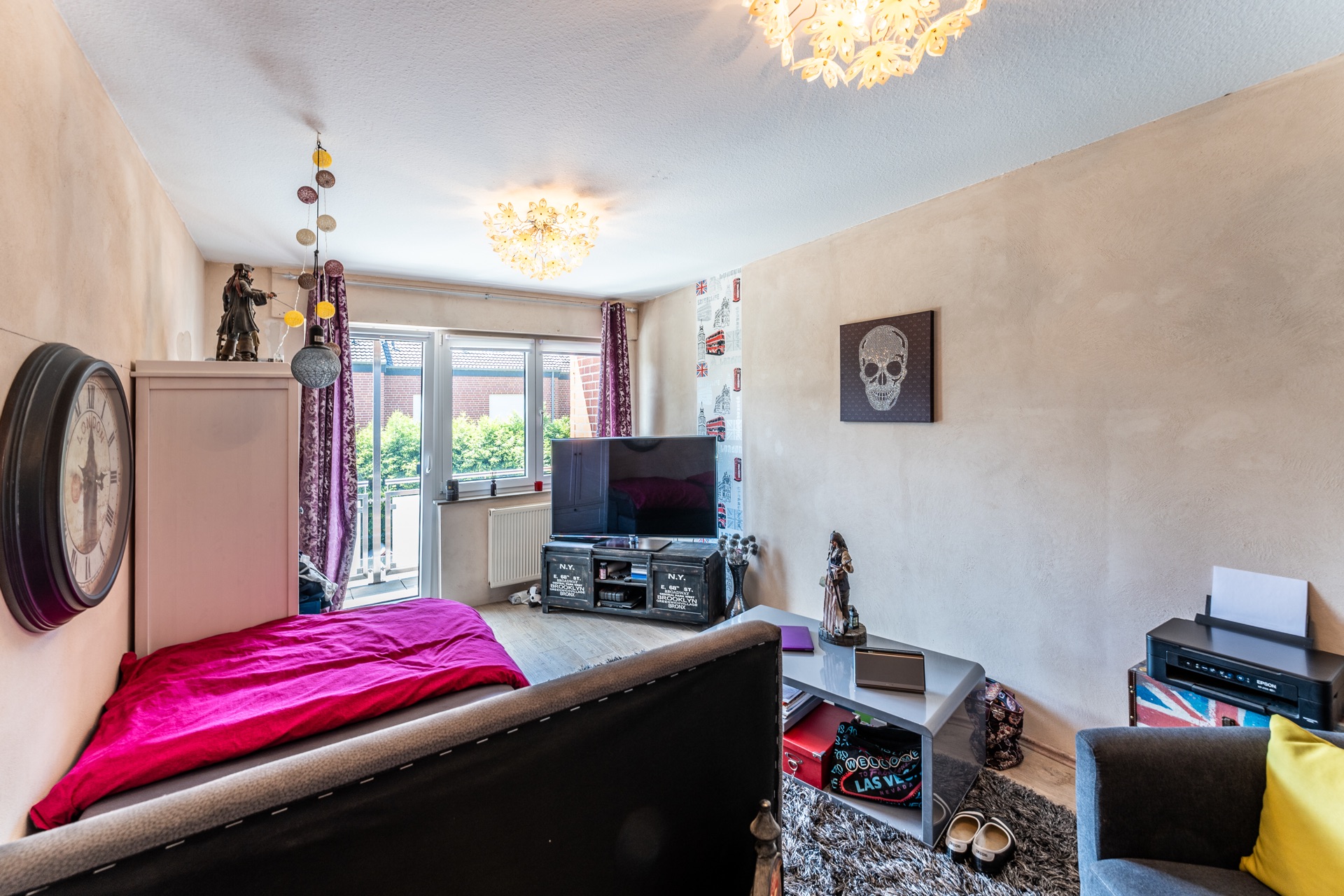 Wohnzimmer mit Sofa, Fernseher, großem Wandgemälde und Zugang zum Balkon.
