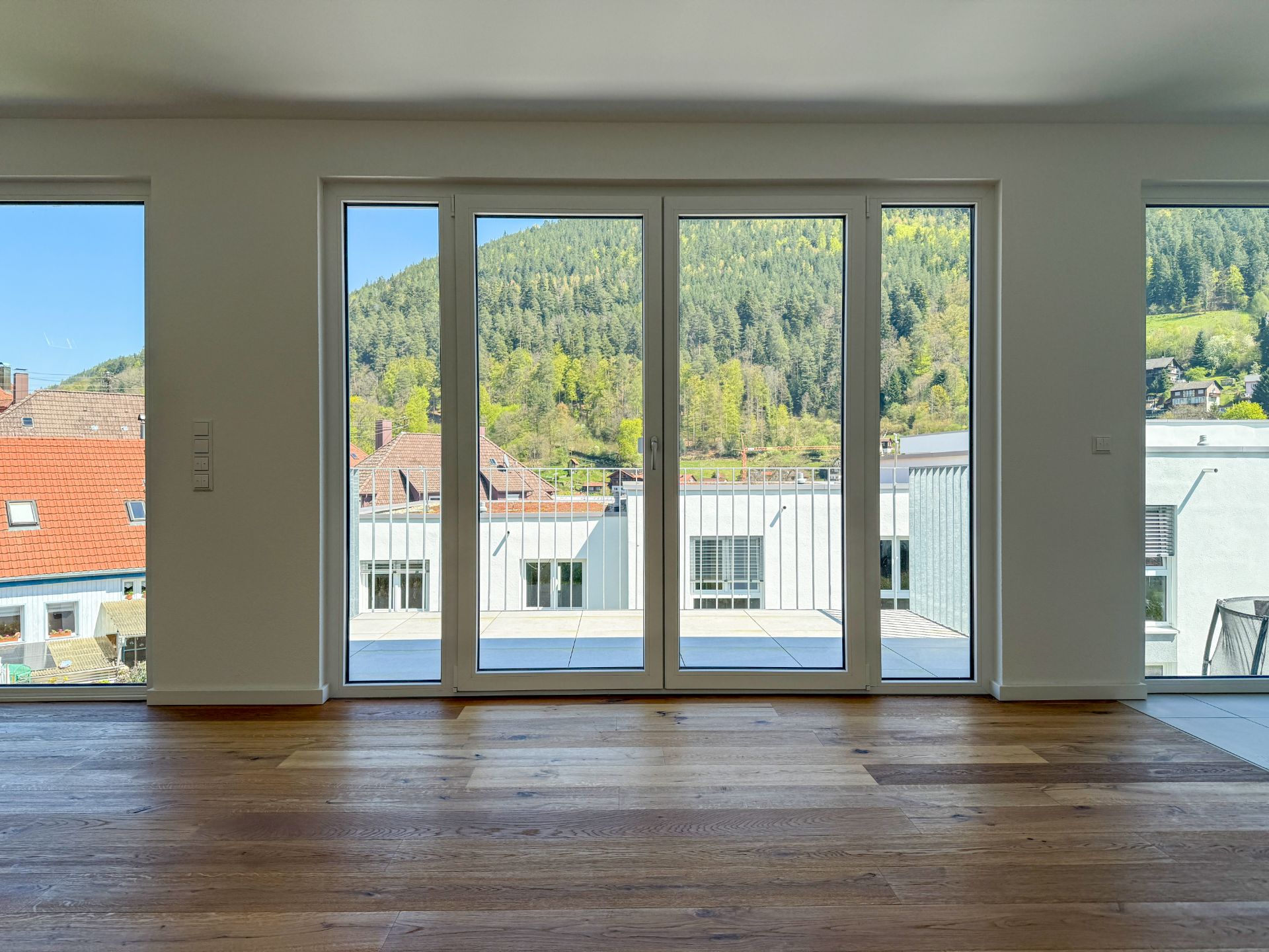 Helles Wohnzimmer mit Parkettboden und großen Fenstern, Blick auf Balkon und grüne Hügel.