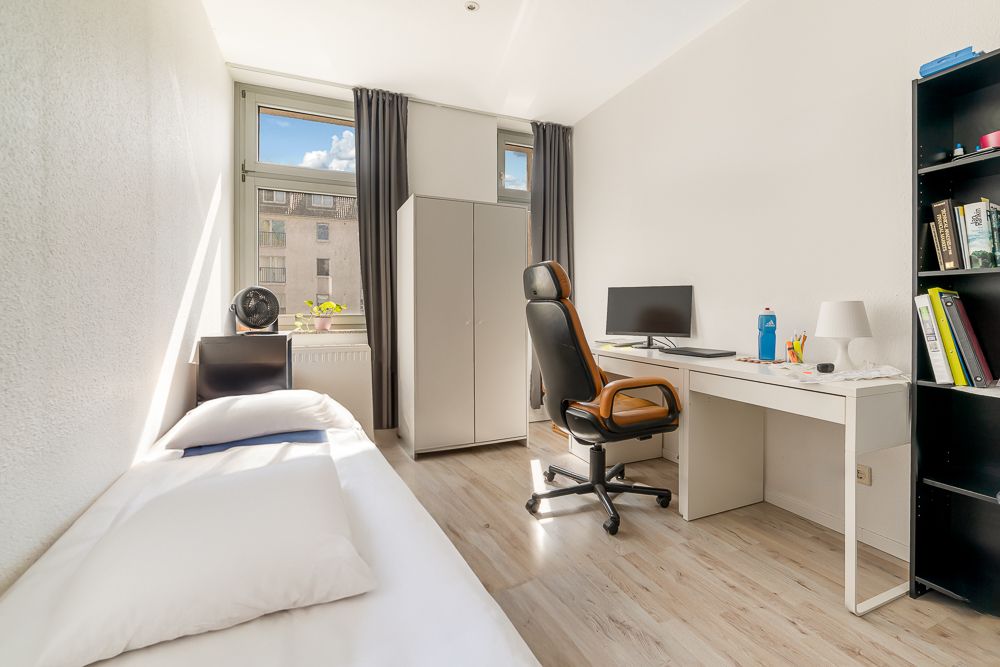 Helles Schlafzimmer mit Einzelbett, Schreibtisch, Stuhl, Schrank und Regal, Fenster im Hintergrund.
