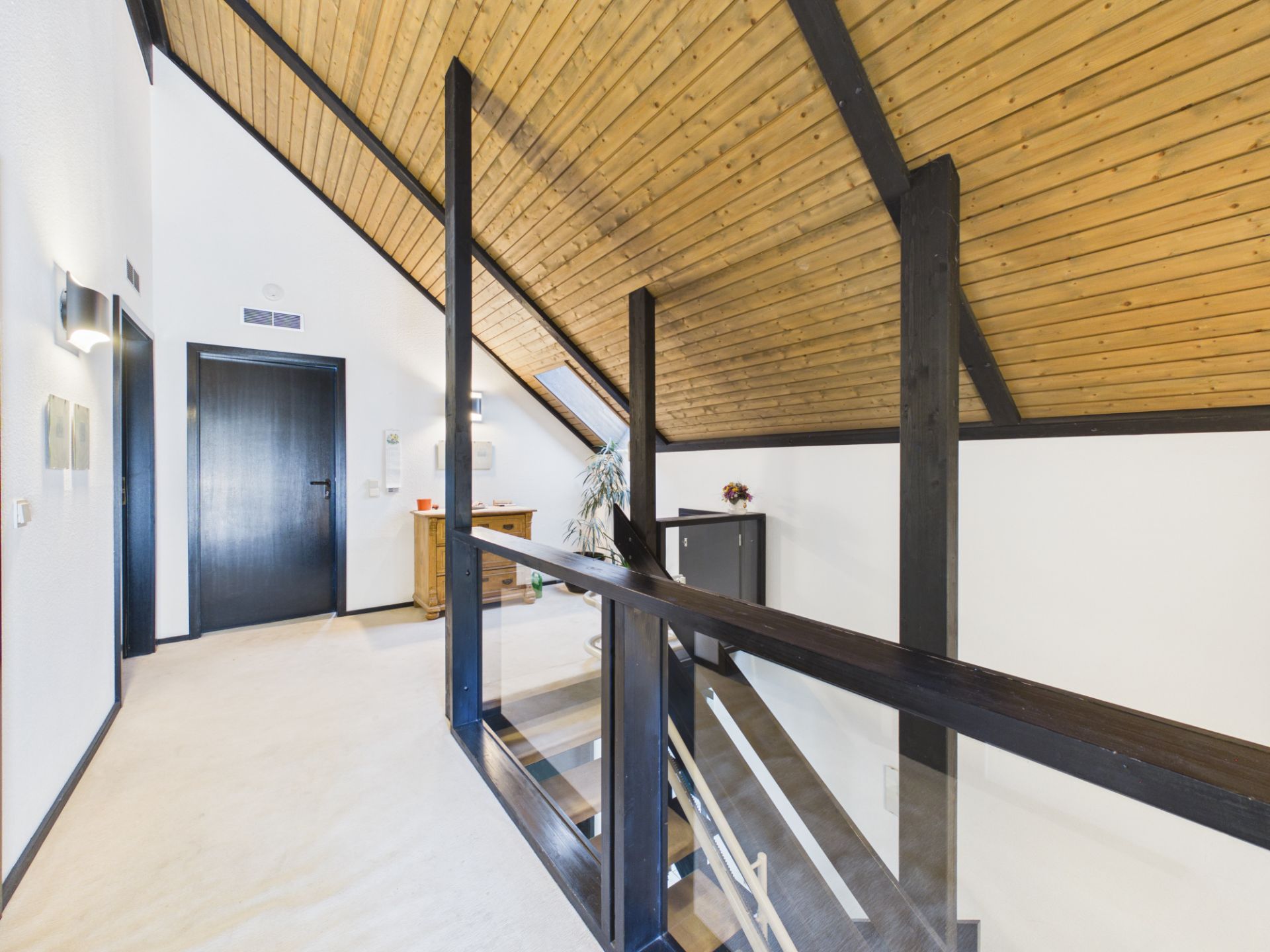 Flur mit schräger Holzdecke, Holztüren, Geländer und Möbelstücken im Obergeschoss.
