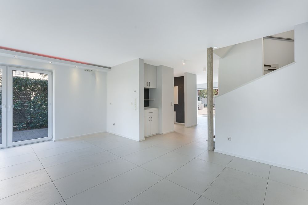 Helles, leeres Wohnzimmer mit Fliesenboden, Glastür zum Garten und offener Treppe.