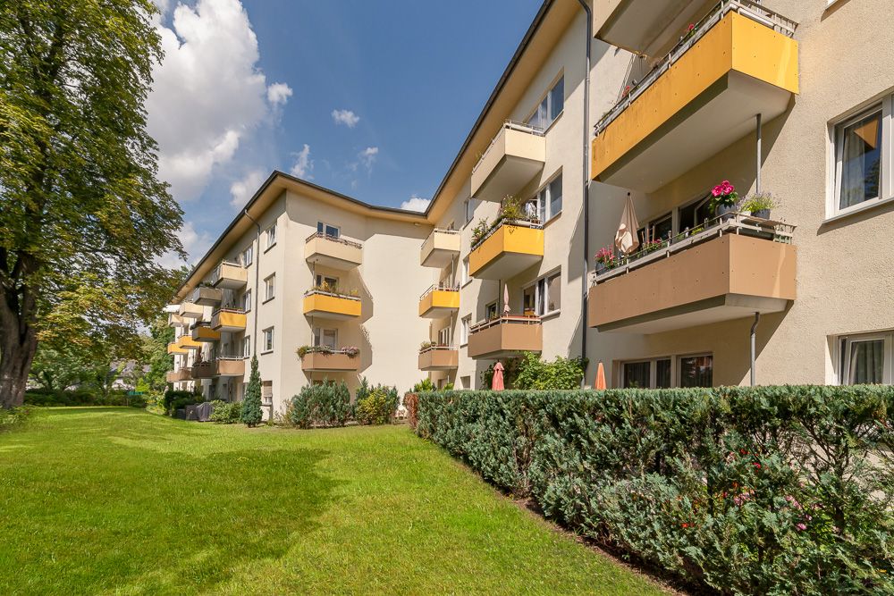 Mehrfamilienhaus mit gelben Balkonen, grünem Rasen und Büschen vor dem Gebäude bei Sonnenschein.