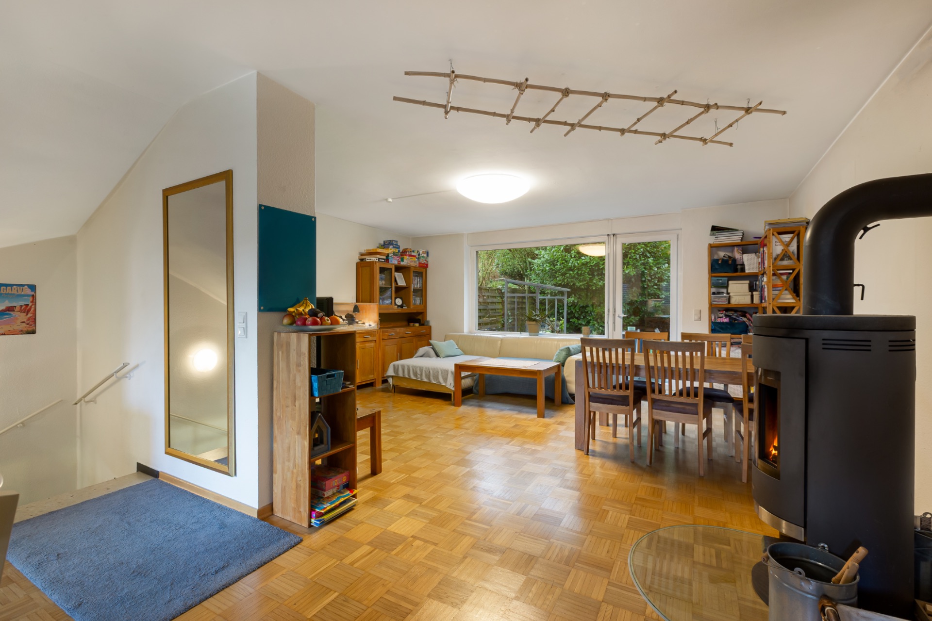 Wohnzimmer mit Holzofen, großem Fenster, Sofa, Esstisch und Holzregalen.