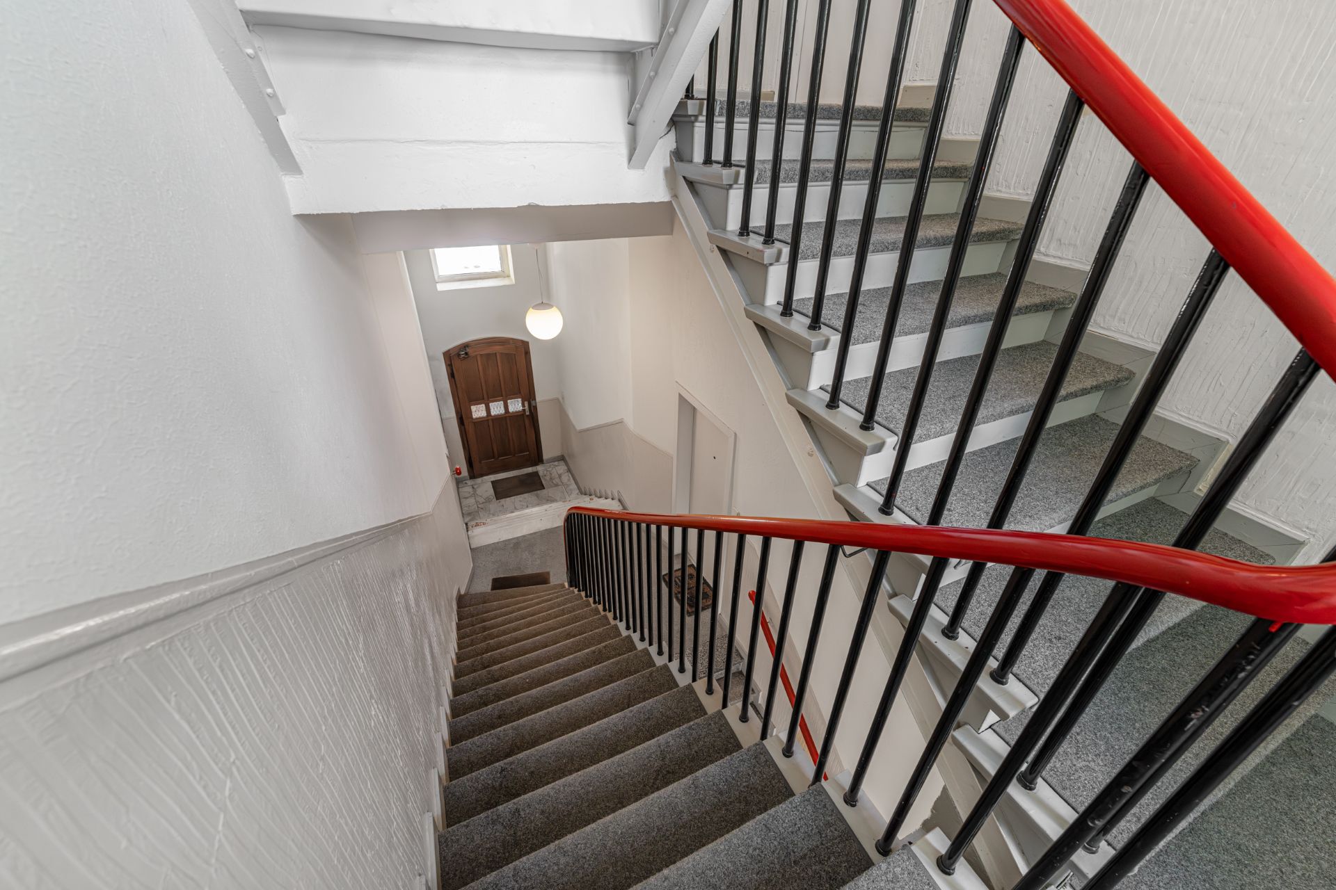 Treppenhaus in einem Wohngebäude mit rotem Handlauf und brauner Eingangstür.
