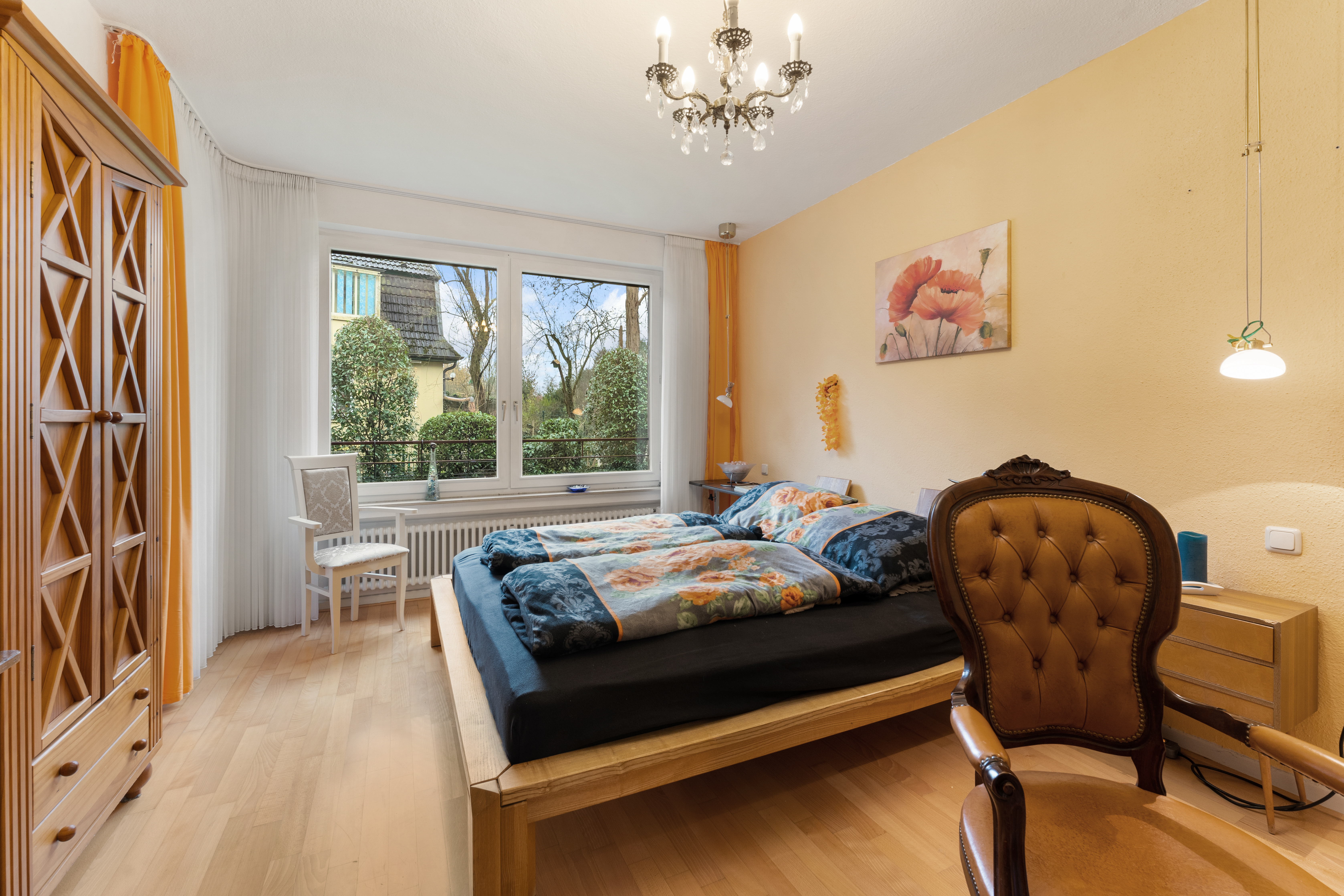 Helles Schlafzimmer mit Doppelbett, großem Fenster, Holzschrank und Stuhl auf Parkettboden.
