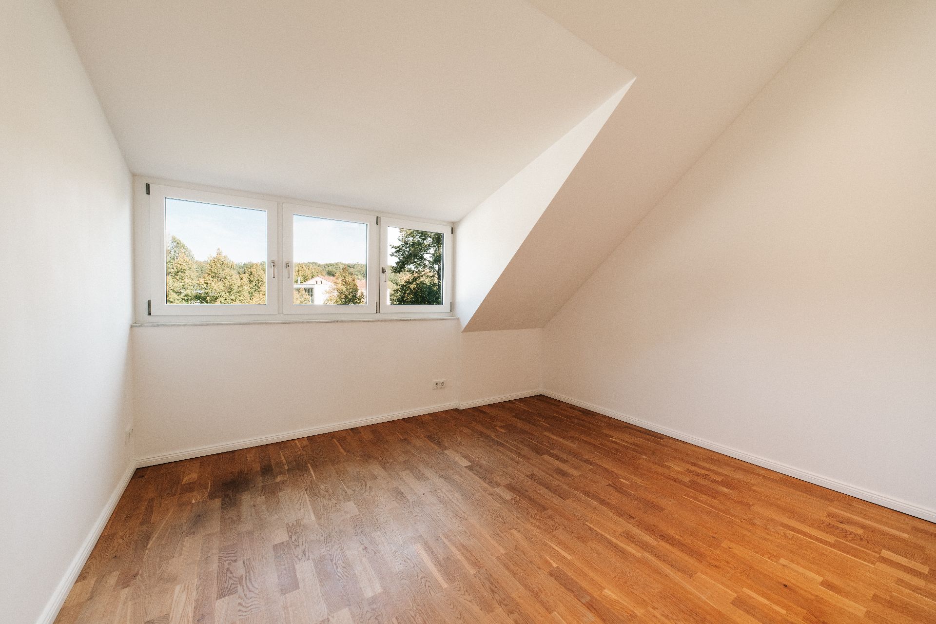 Leeres Zimmer mit Holzfußboden und drei Fenstern unter einer Dachschräge.