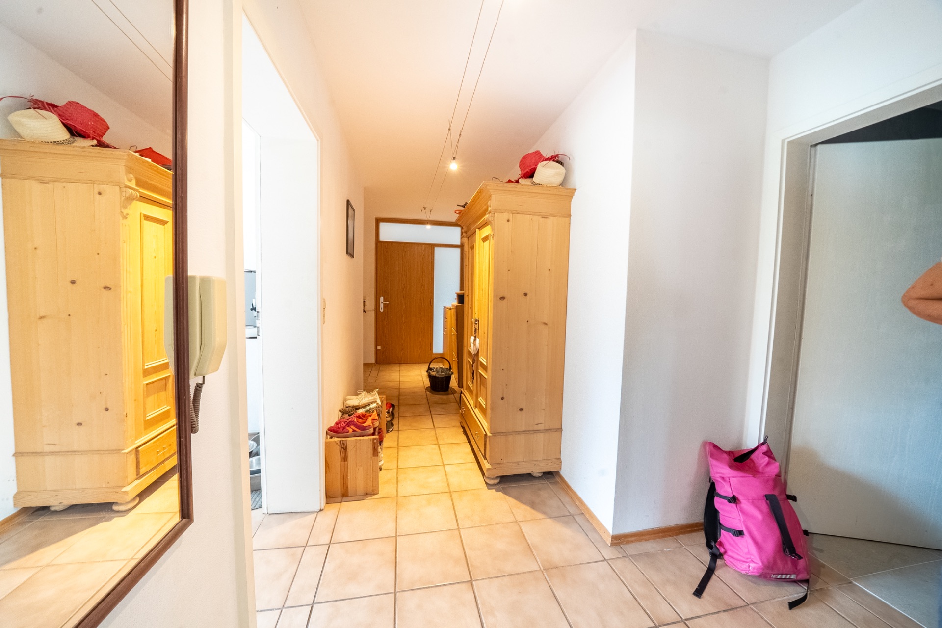 Heller Flur mit zwei Holzschränken, Spiegel, Kisten und einer rosa Tasche auf Fliesenboden.