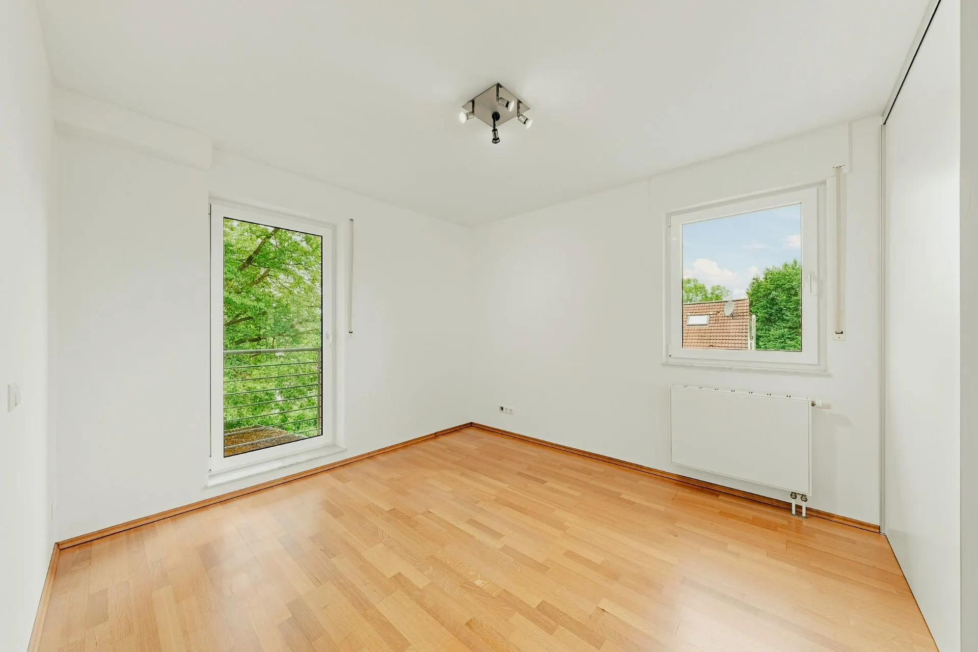 Leerer Raum mit Holzfußboden, weißen Wänden, Fenster und Glastür mit Blick ins Grüne.