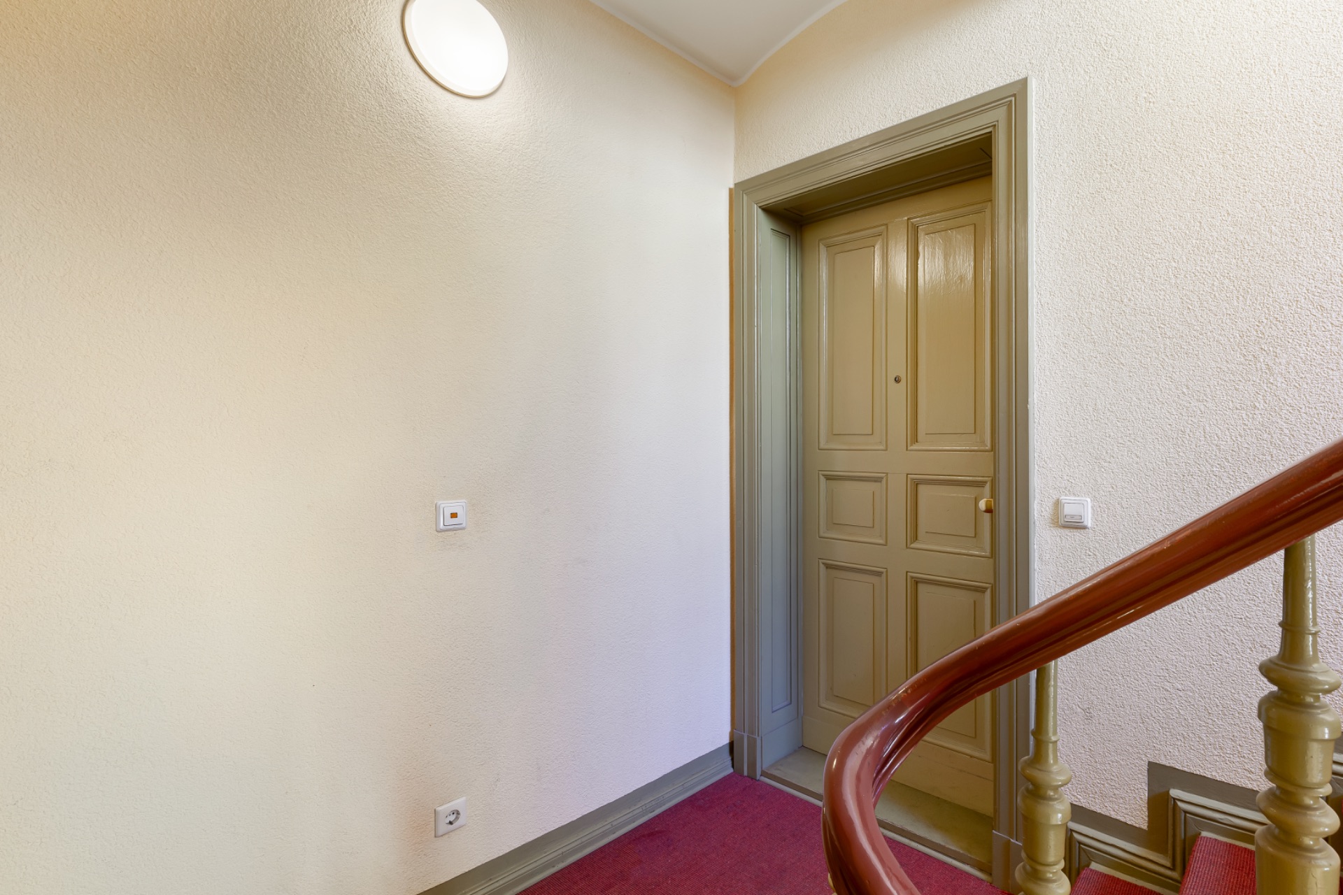 Treppenhaus mit rotem Teppich und beiger Wohnungstür.