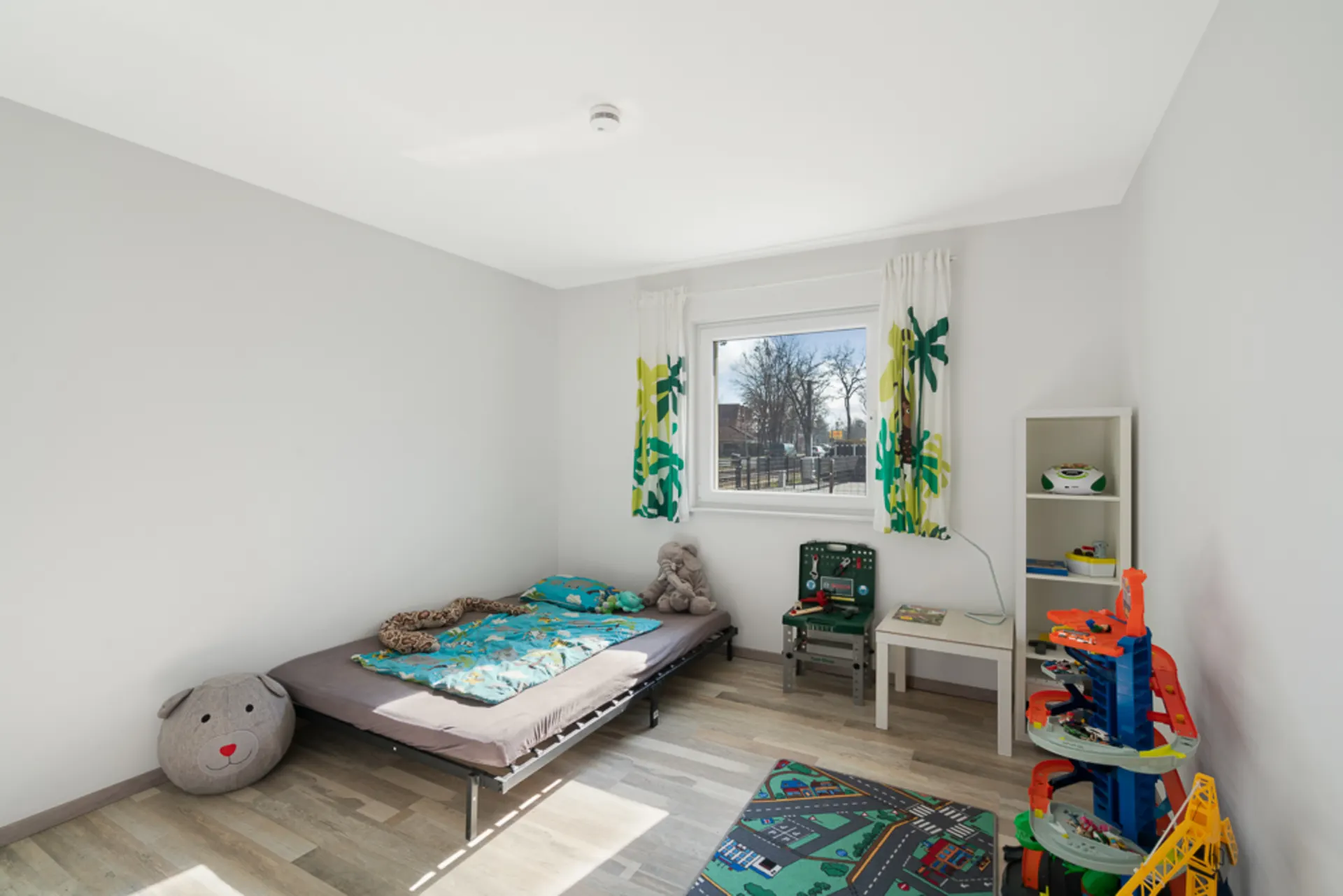 Helles Kinderzimmer mit Bett, Spielzeug, Regal und Fenster mit gemusterten Vorhängen.