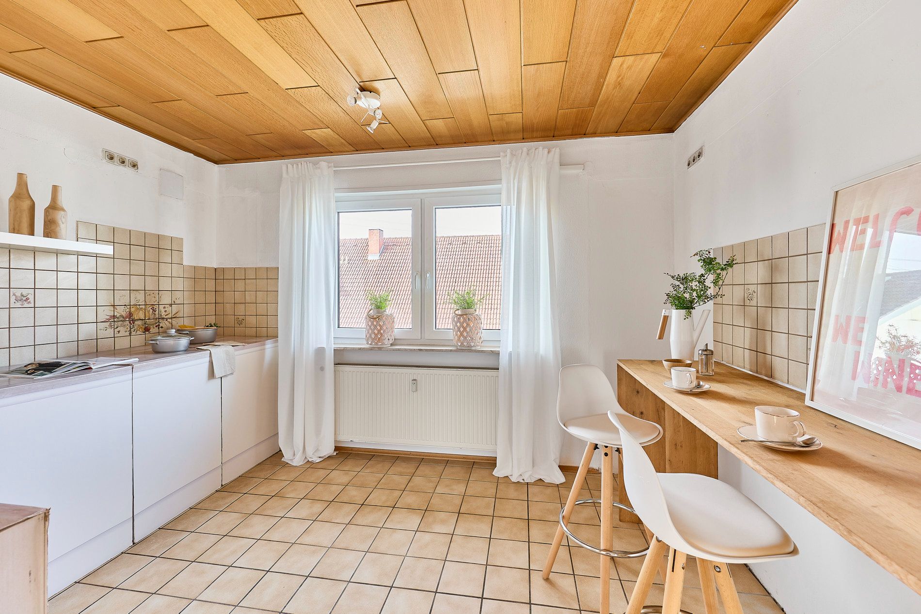 Helle Küche mit Holzdecke, Fliesenboden, Bartresen und zwei weißen Barhockern.