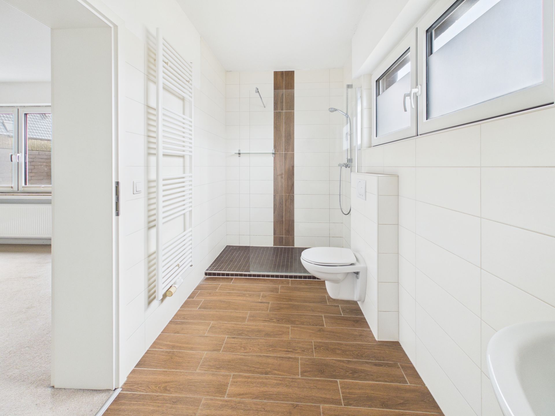 Helles Bad mit Holzoptik-Boden, begehbarer Dusche, Wandtoilette und zwei Fenstern.