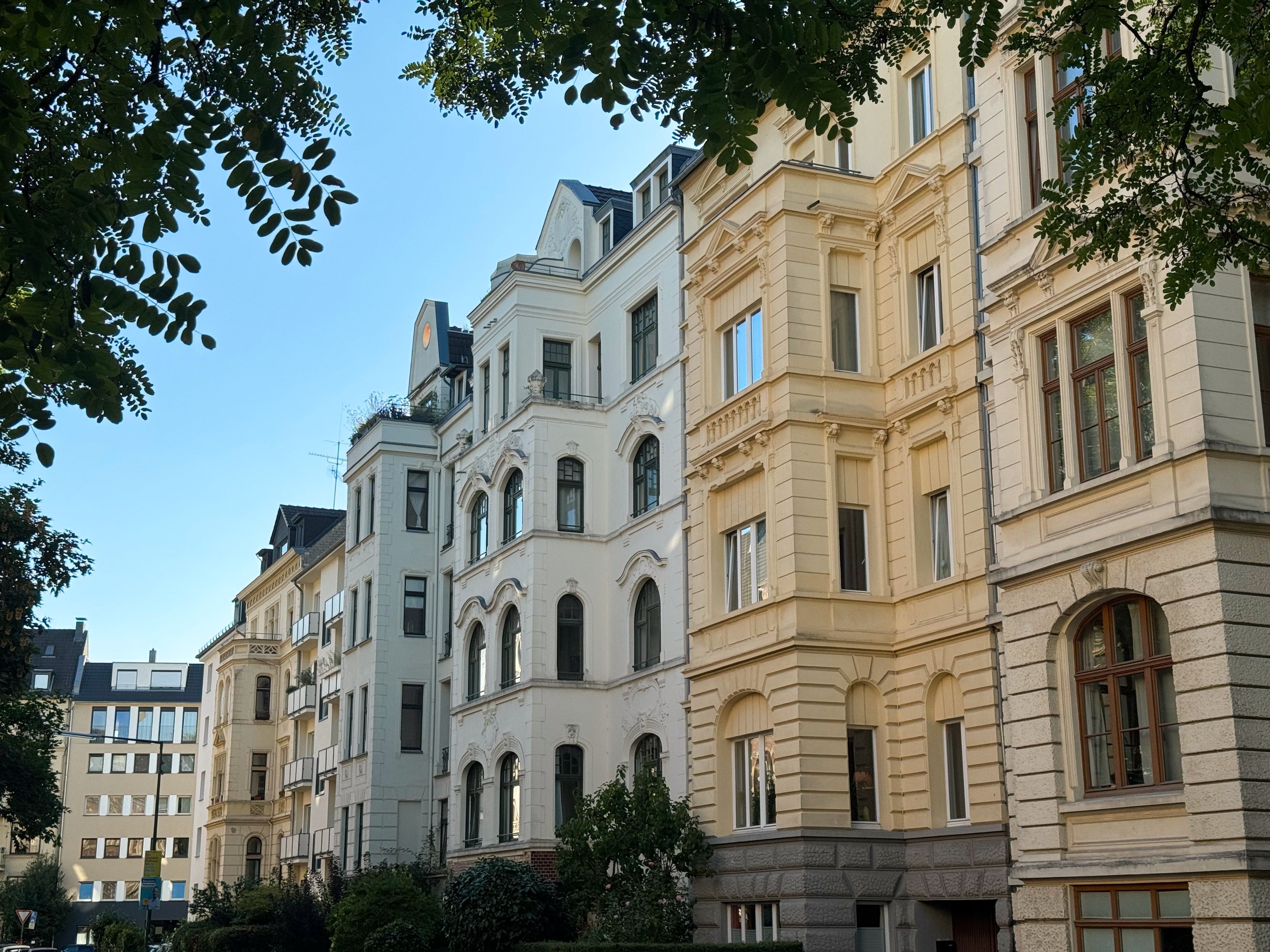 Mehrstöckige Wohnhäuser mit klassischen Fassaden in einer ruhigen Straße, teils von Bäumen beschattet.