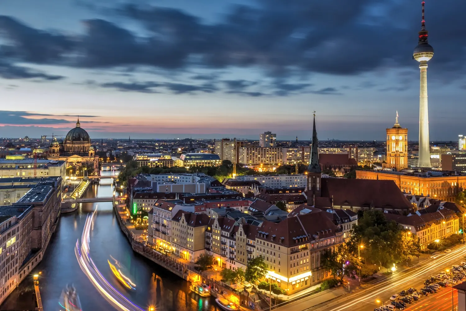 Abendliche Stadtansicht mit Fluss, beleuchteten Gebäuden und Fernsehturm im Hintergrund.
