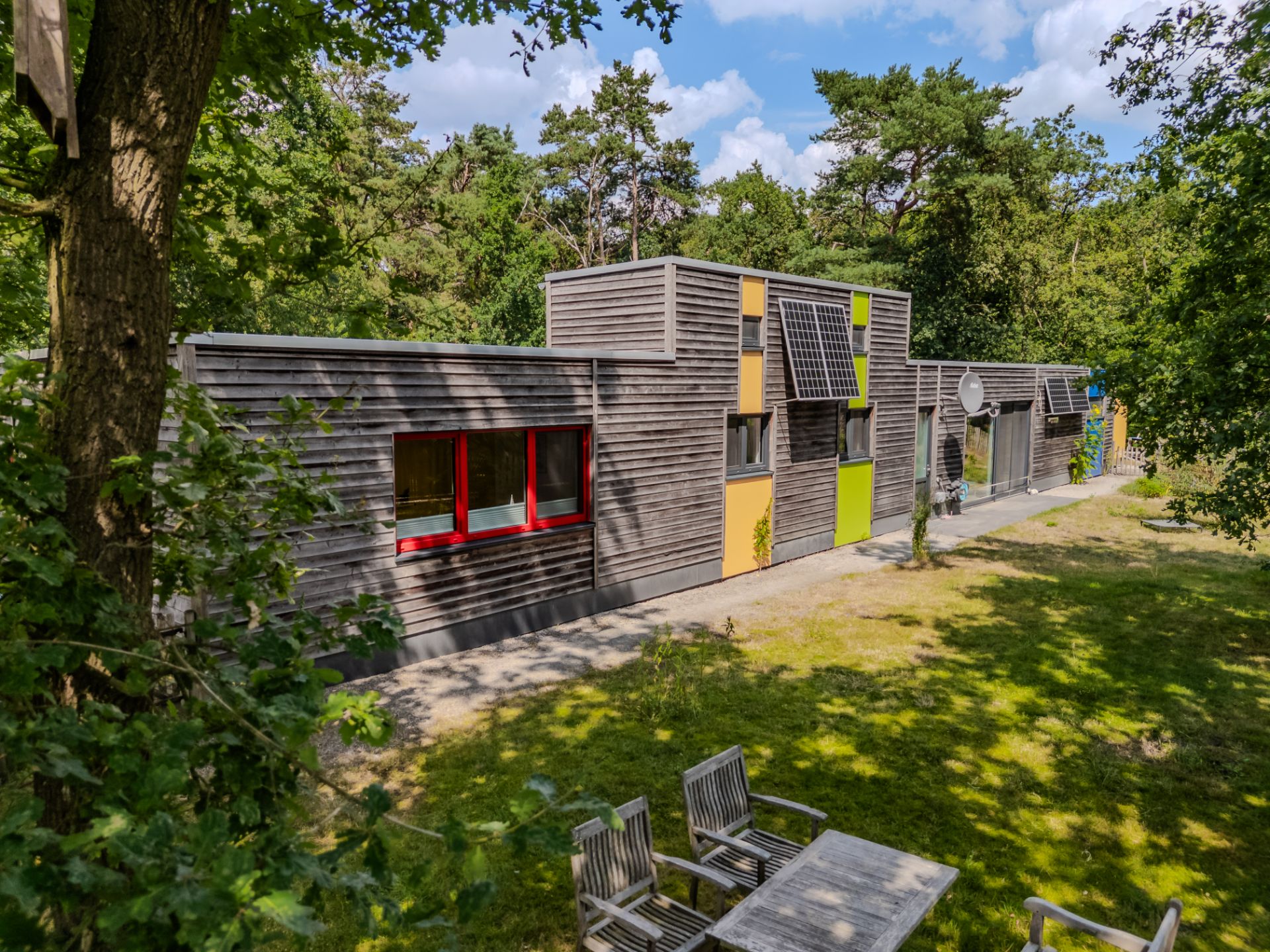 Modernes Holzhaus mit bunten Akzenten und Terrassenmöbeln im Garten, umgeben von Bäumen.