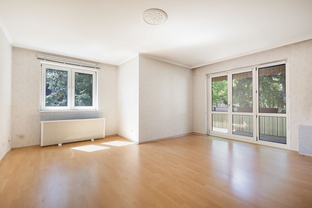 Helles, leeres Zimmer mit Parkettboden, Fenster, Heizung und Zugang zum Balkon.