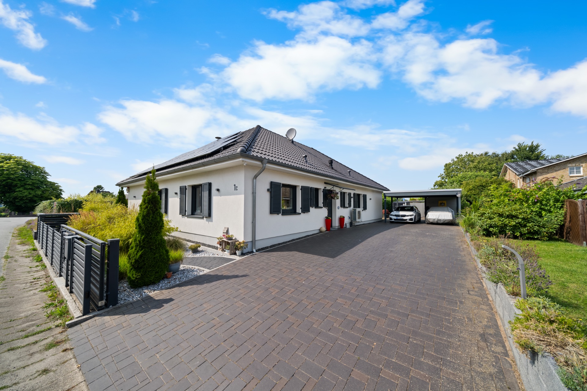 Einfamilienhaus mit dunklem Dach, gepflasterter Einfahrt und Carport mit geparktem Auto.