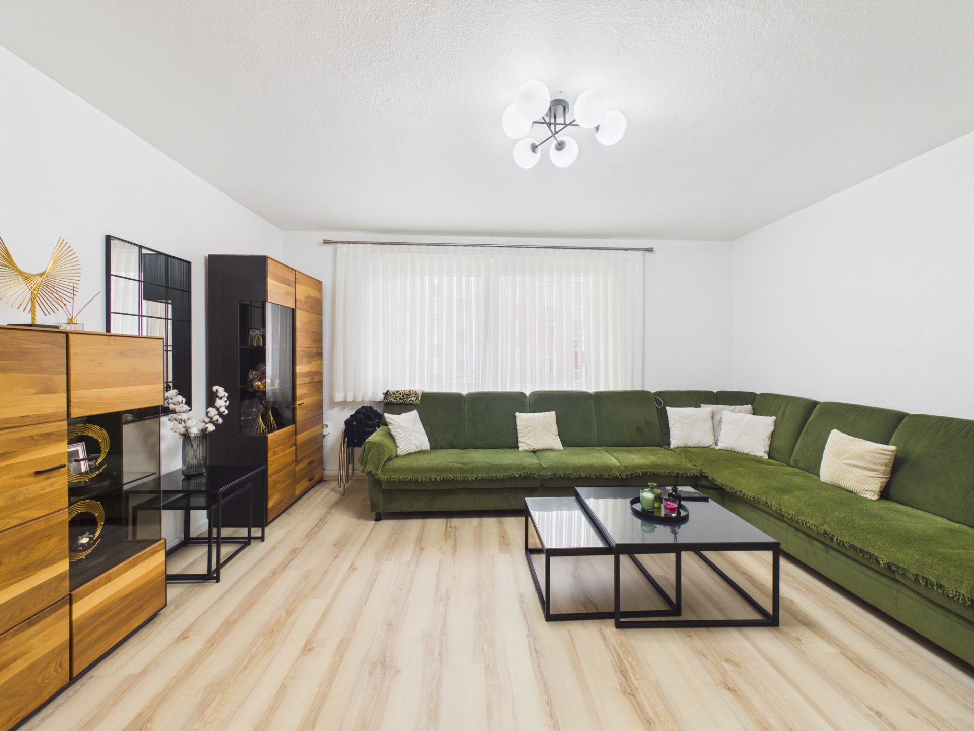 Wohnzimmer mit großer grüner Eckcouch, Holzregalen und zwei Couchtischen auf hellem Boden.