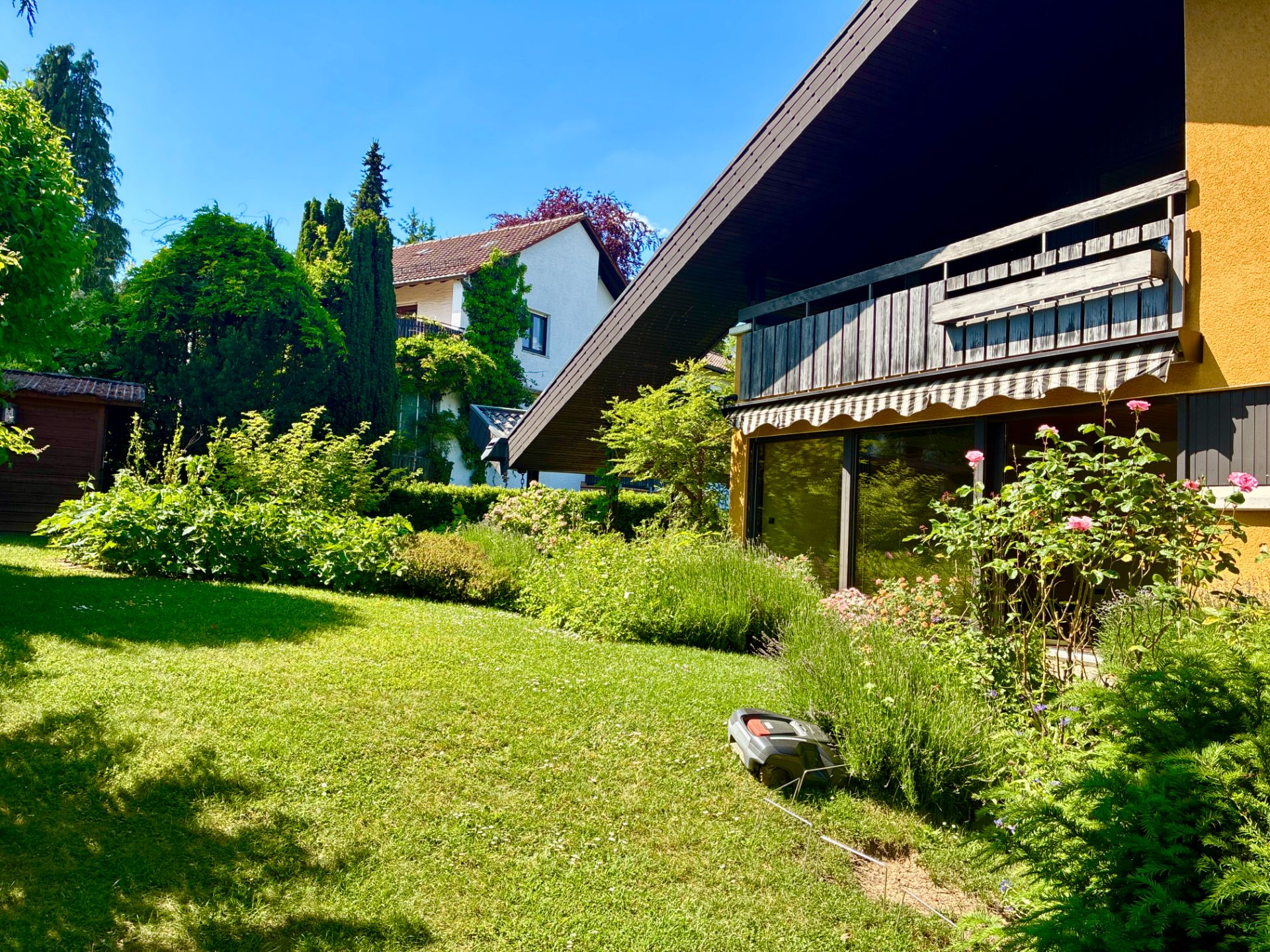 Garten mit Rasenfläche, Blumenbeeten und einem Haus mit großer Terrasse im Hintergrund.
