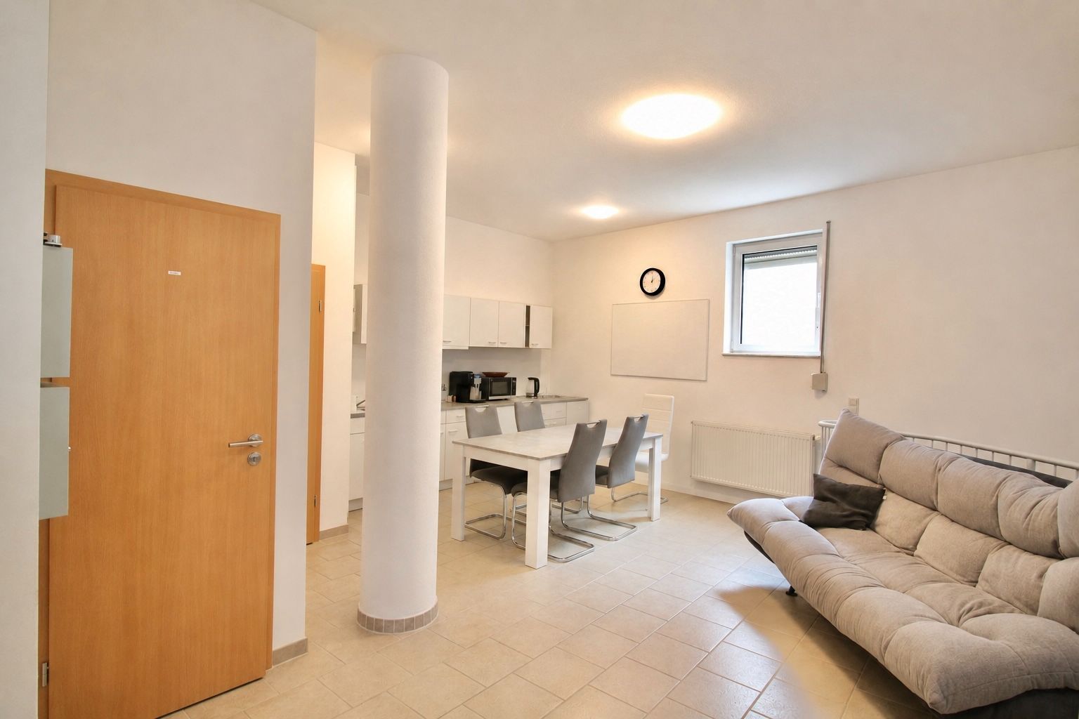 Helles Wohnzimmer mit Sofa, Esstisch, vier Stühlen, Fenster und Küche im Hintergrund.