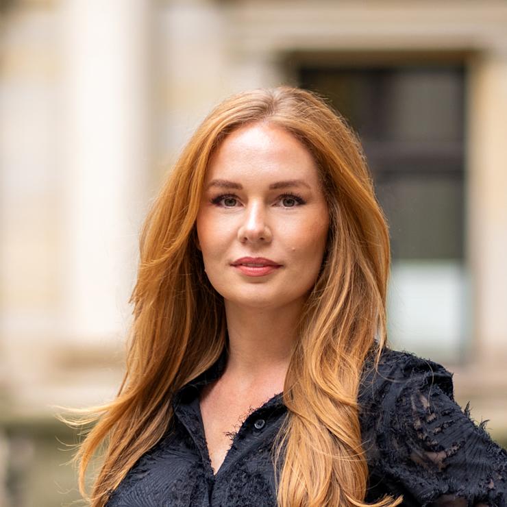 Frau mit langen roten Haaren und schwarzer Bluse steht draußen vor einem Gebäude.