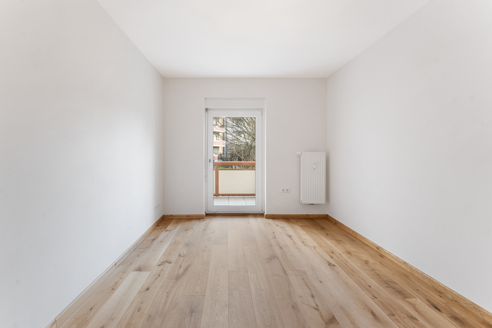 Heller, leerer Raum mit Holzboden und Zugang zu einem Balkon durch eine Glastür.