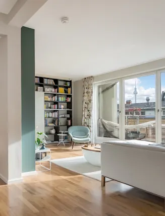 Helles Wohnzimmer mit Holzboden, Sessel, Regal, Sofa und großem Fenster zur Terrasse.